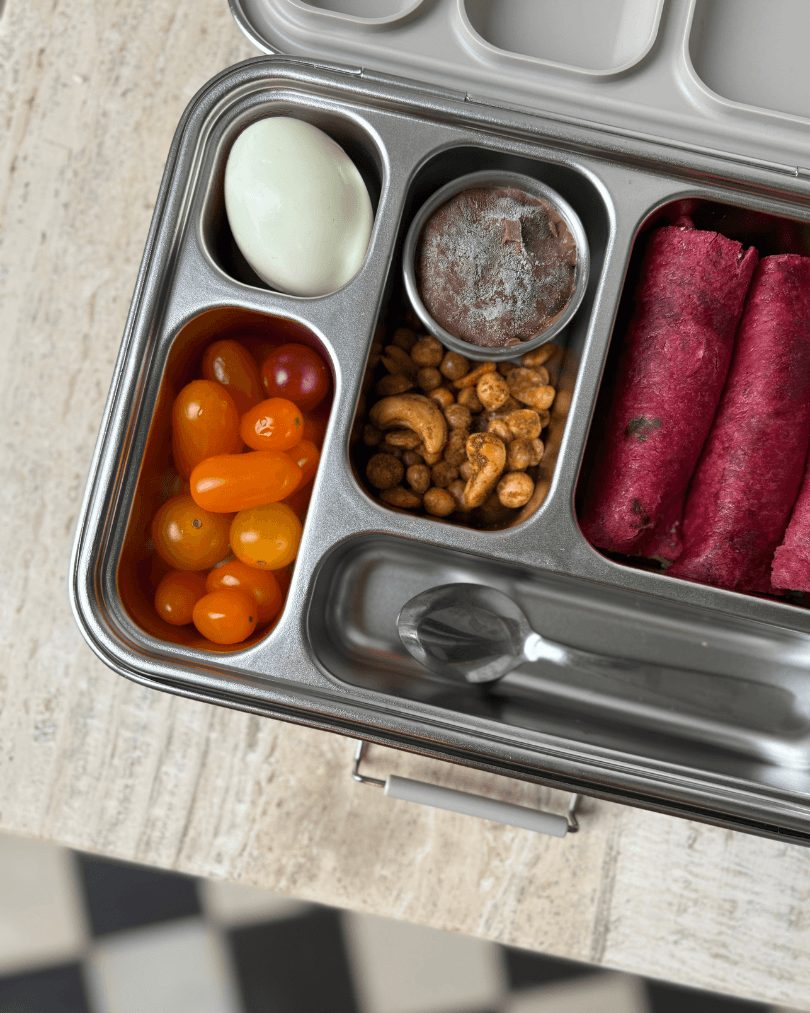 An open stainless lunchbox packed with boiled egg, tomatoes, spiced nuts, beetroot wraps and a chocolate treat