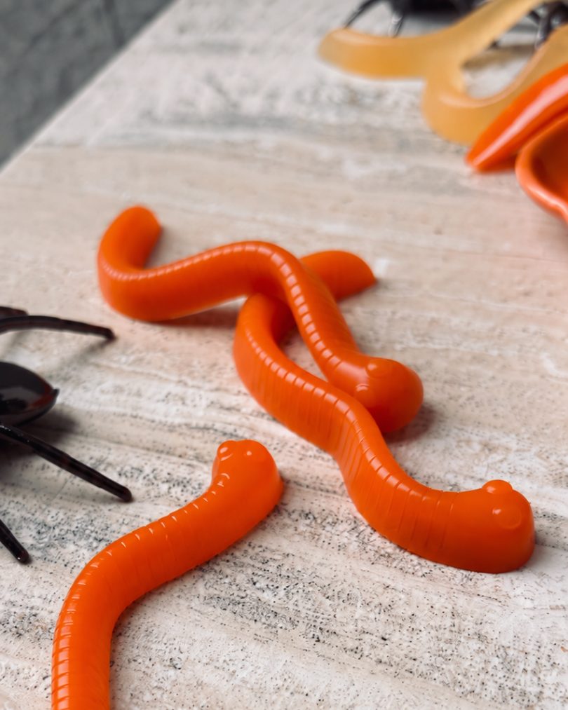 Bright orange mango carrot gummy worms on a pale textured surface beside Halloween spider decorations and silicone worm moulds.