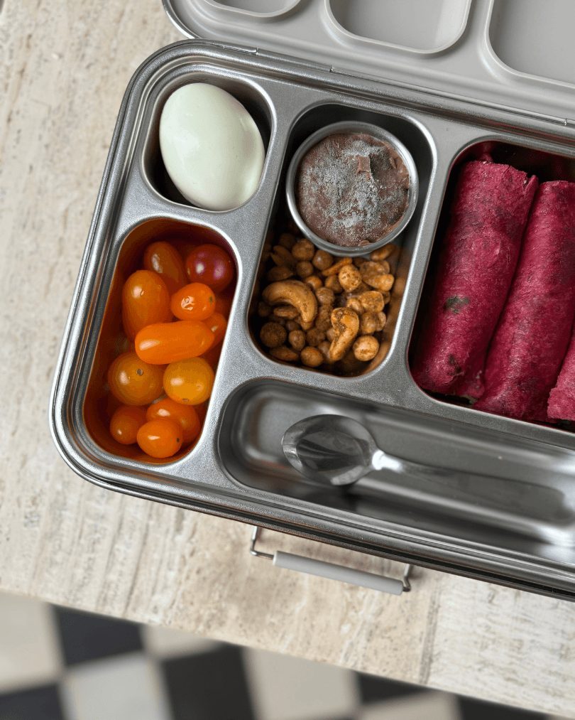 Open stainless-steel lunchbox packed with an egg, cherry tomatoes, chocolate dip, crunchy snacks and beetroot wraps