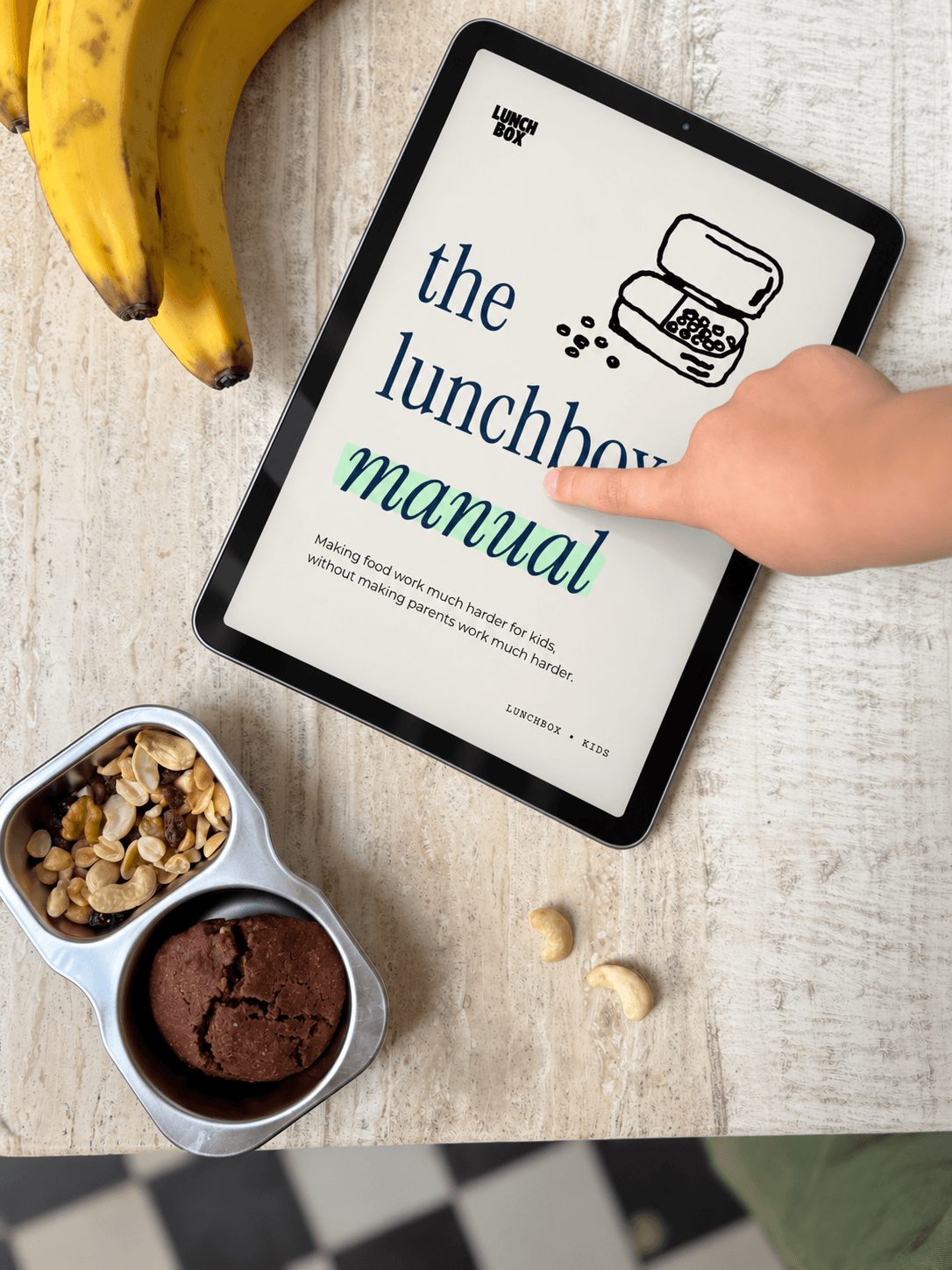 The Lunchbox Manual on a tablet beside a packed stainless snack tin and bananas