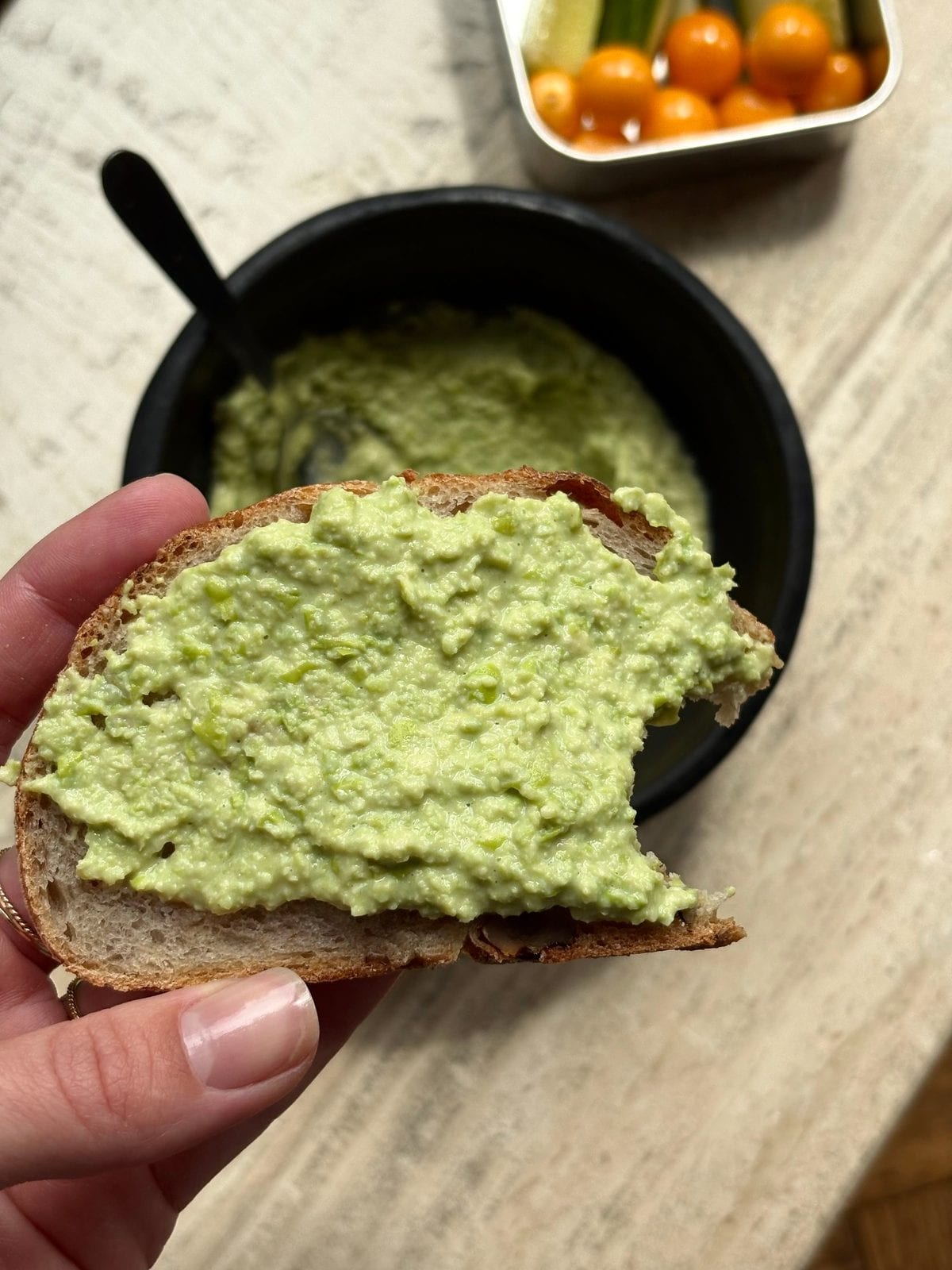 A slice of sourdough bread thickly spread with creamy green edamame dip, held over a small black bowl with more dip and a lunchbox of cherry tomatoes and cucumber sticks in the background.