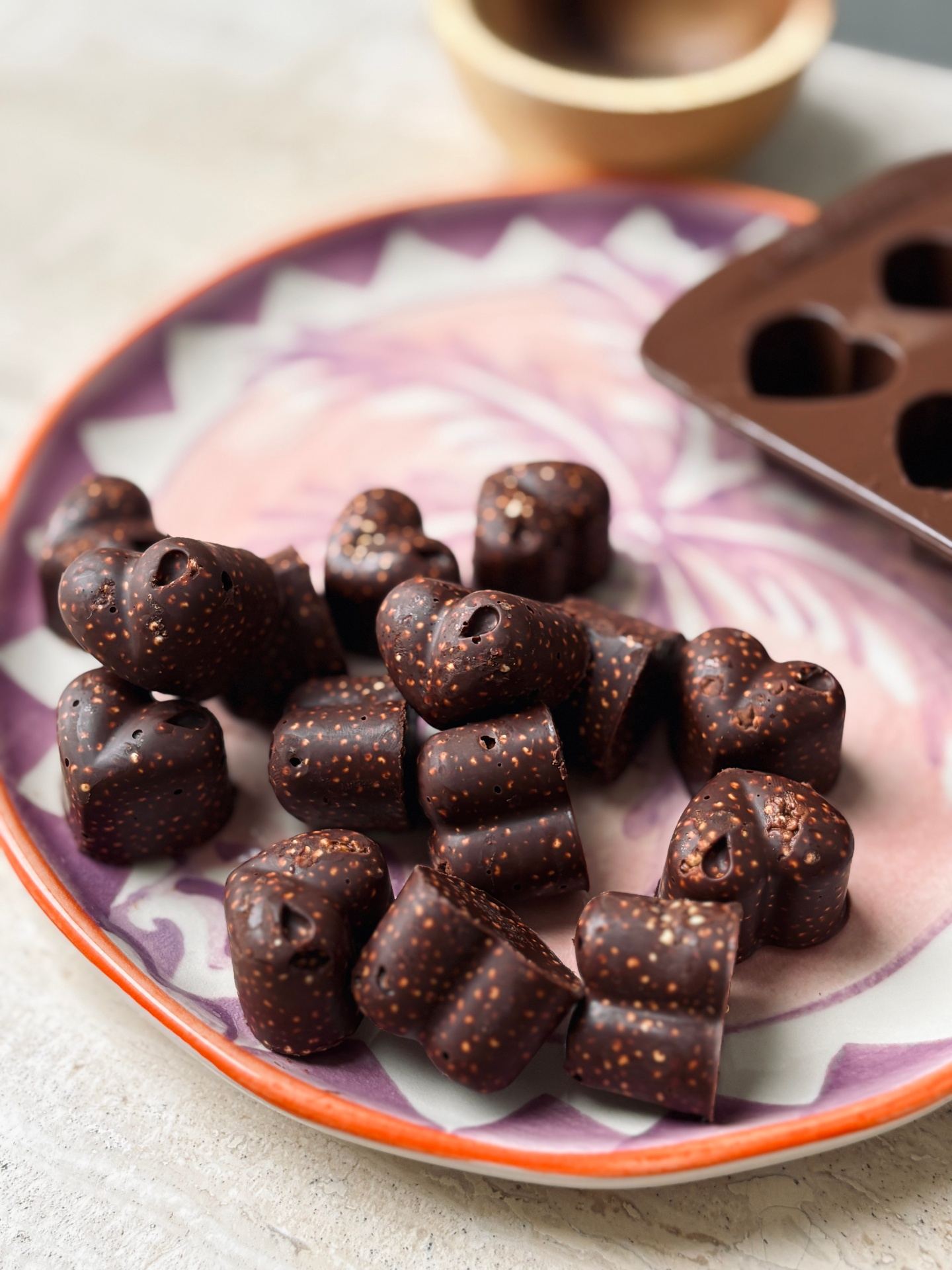 A pile of small dark chocolate pieces speckled with popped quinoa on a pink and white patterned plate with a heart-shaped silicone chocolate mould in the background.