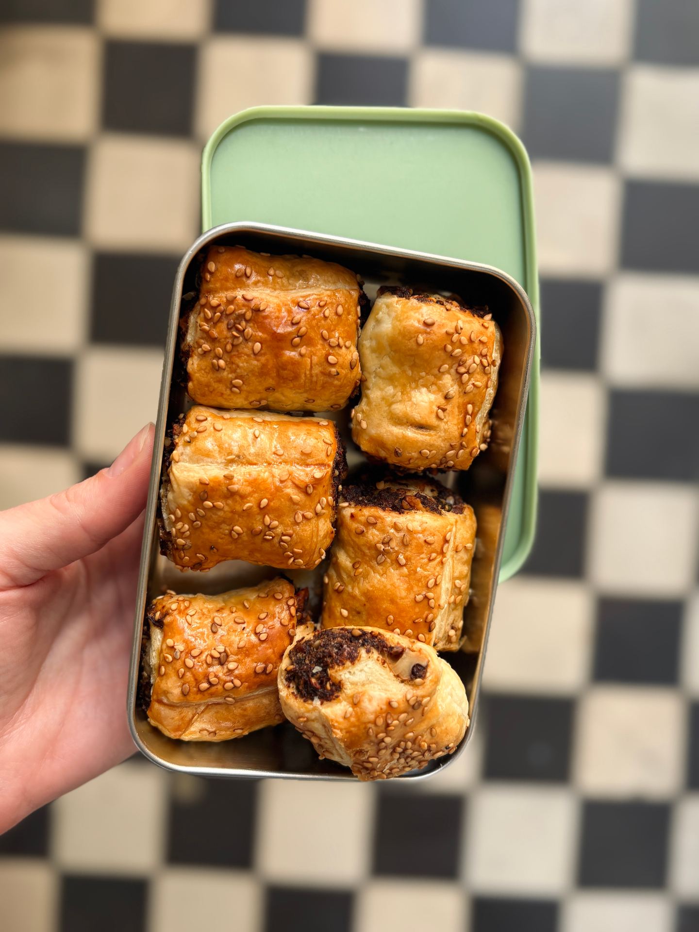 A hand holding a green-lidded lunchbox tin packed with golden, sesame-topped puff pastry mushroom rolls over a black-and-white checkered background.