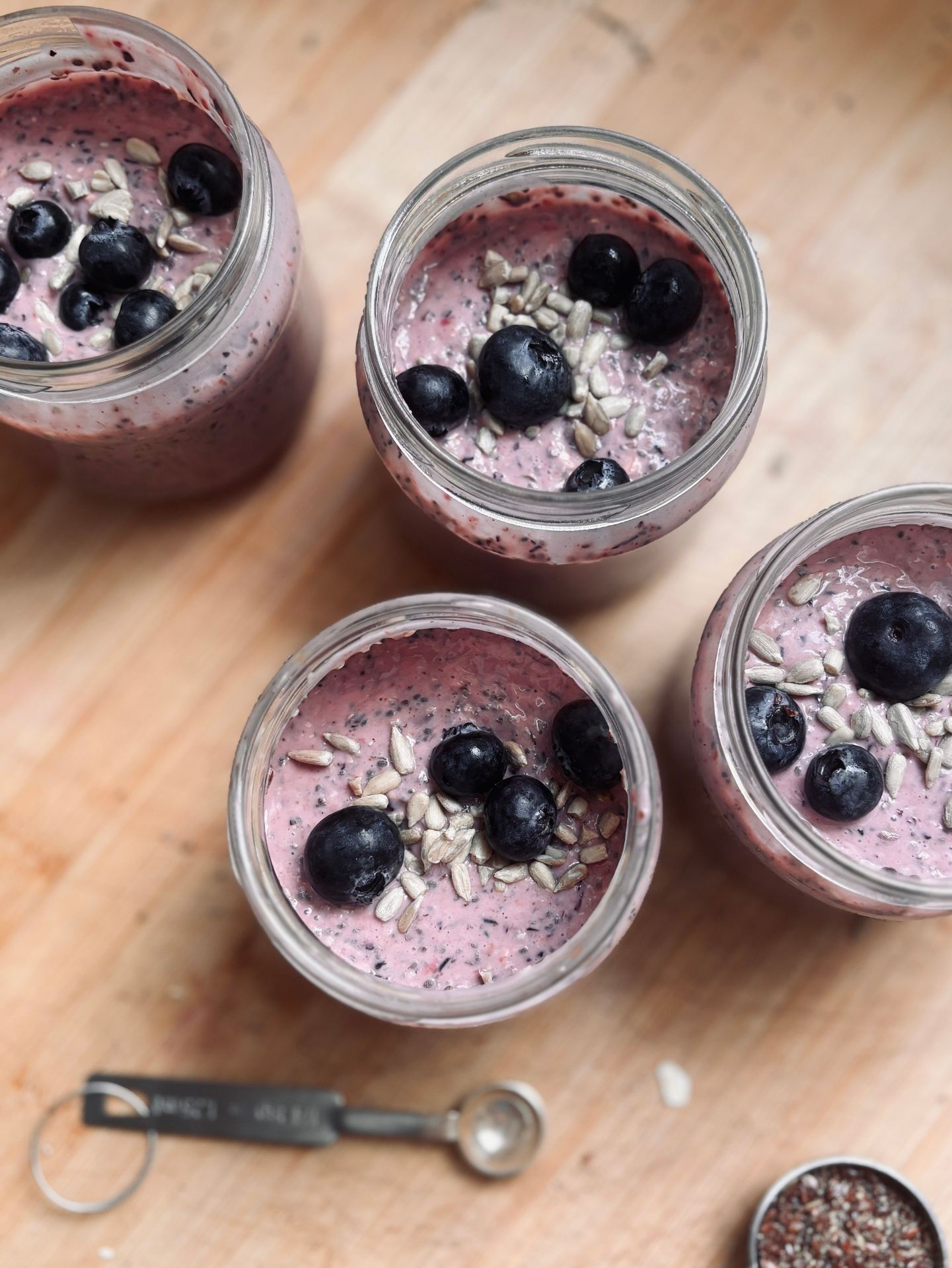 Four small glass jars of creamy pink berry chia pudding topped with fresh blueberries and sunflower seeds, sitting on a wooden board next to a measuring spoon and a bowl of chia seeds.