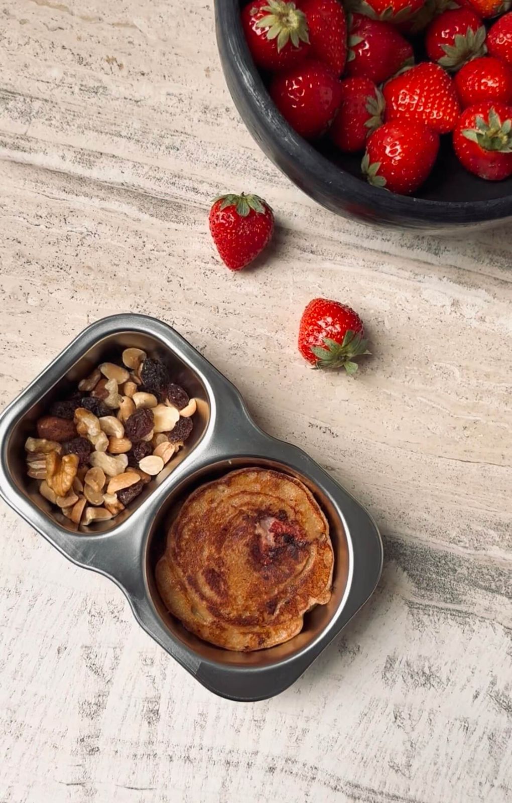 Two-compartment stainless steel lunchbox holding a golden strawberry protein pancake and a trail mix of nuts and raisins, with fresh strawberries beside it.